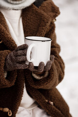 Cropped photo of little girl in warm clothes and mittens holding a big white metal cup of hot drink in hands in snowy forest. Adventure with family on picnic on winter holiday vacation