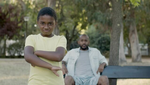 Young black boy posing at camera, folding arms and frowning. Blurred image of father sitting on bench in park in background. Front view. Medium shot. Father and son, generations concept.