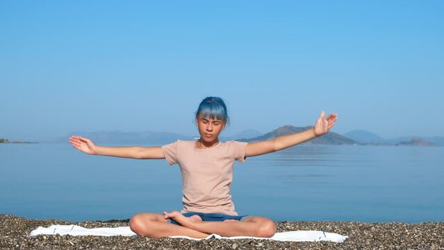 Girl stretching hands yoga on sunny shore. A young girl stretching her hands and relax in lotus pose on the beach against sea view.