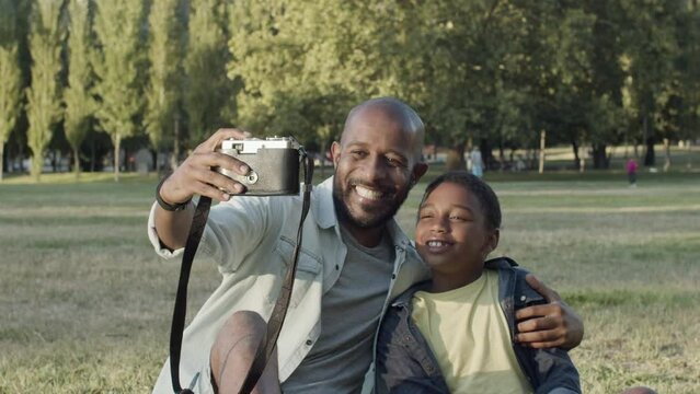 Father and son taking selfie in park, making funny faces and smiling. Black bearded man holding camera while playing the ape with his son and showing tongues. Father and son, happy family concept.