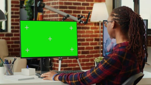 African american woman looking at computer with greenscreen template, working with blank copyspace chromakey. Analyzing pc desktop with isolated mockup background on monitor in office space.