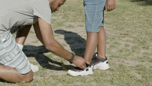 Black father tying his young sons shoelaces in summer park. Parent and child spending time together outside on sunny day, having stopped for fixing laces on sons snickers. Parenthood, family concept.