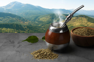 Calabash with mate tea and bombilla on light grey table and beautiful view of mountain landscape,...