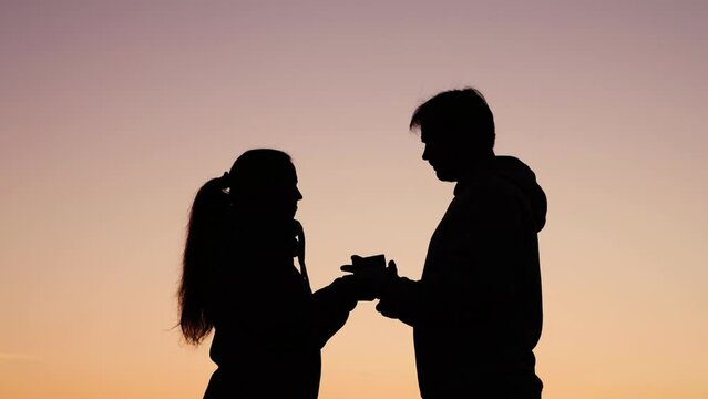 Young guy gives box with surprise to his girlfriend, happy couple hugs, gentle kiss in nature. Valentines Day. Silhouette man gives gift to his beloved woman outdoors against sky. Romantic love, park