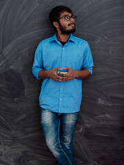 Indian smiling young student in blue shirt and glasses using smartphone and posing on school blackboard background