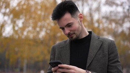 Happy young handsome Caucasian guy standing on autumn forest messaging in smartphone app smiling. Portrait of confident brunette man enjoying overcast weather in park scrolling social media