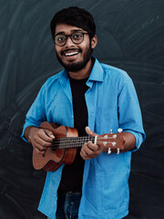 Indian young man in a blue shirt and glasses playing the guitar in front of the school blackboard