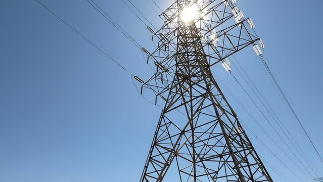 Large electrical tower and power lines against blue sky and bright sun, tilt up; in Ontario Canada.