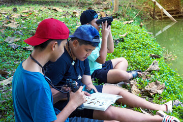 Asian boy and friends Invite each other to see birds in the community forest on holidays. And help each other to search for the types of birds found from the birds Guide book. - obrazy, fototapety, plakaty
