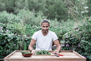 caucasian guy smiling at camera behind a wooden table with marijuana plants on it surrounded by plants in a garden