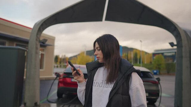 Woman with an electirc vehicle charging and using phone