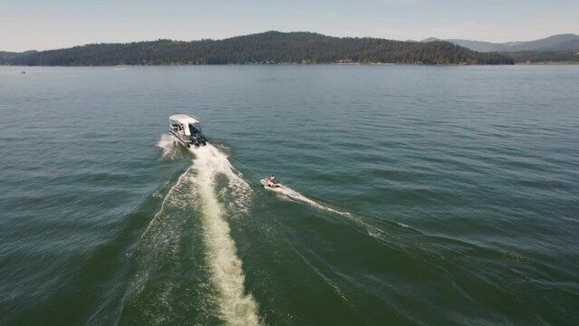 Pontoon boat pulling two kids on a tube on a lake with mountains