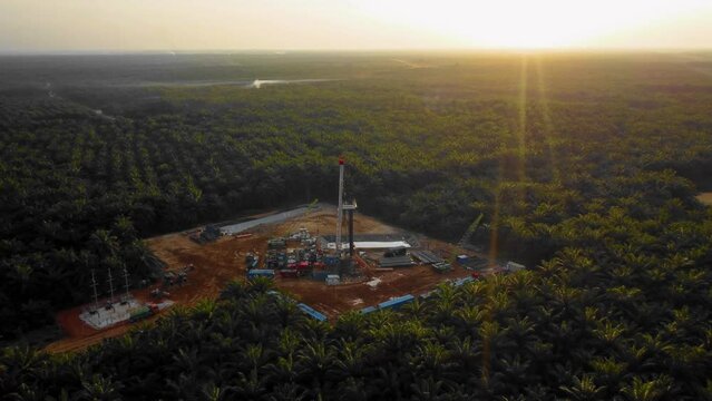 Cinematic 4K Drone Footage of Onshore Drilling Rig equipment structure for oil exploration and exploitation in the middle of jungle surrounded by palm oil trees during sunset and high oil price.