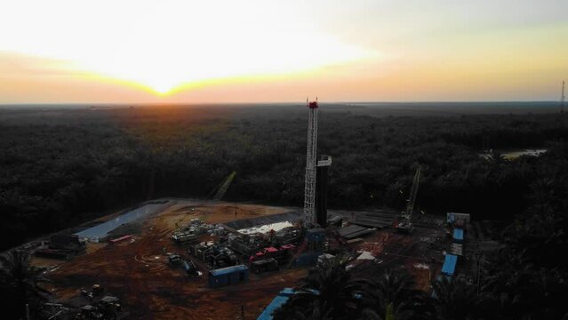 Cinematic 4K Drone Footage of Onshore Drilling Rig equipment structure for oil exploration and exploitation in the middle of jungle surrounded by palm oil trees during sunset and high oil price.