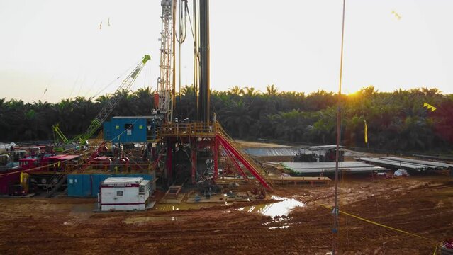 Cinematic 4K Drone Footage of Onshore Drilling Rig equipment structure for oil exploration and exploitation in the middle of jungle surrounded by palm oil trees during sunset and high oil price.