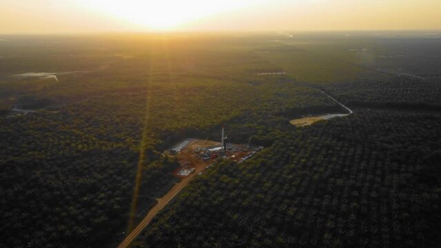 Cinematic 4K Drone Footage of Onshore Drilling Rig equipment structure for oil exploration and exploitation in the middle of jungle surrounded by palm oil trees during sunset and high oil price.
