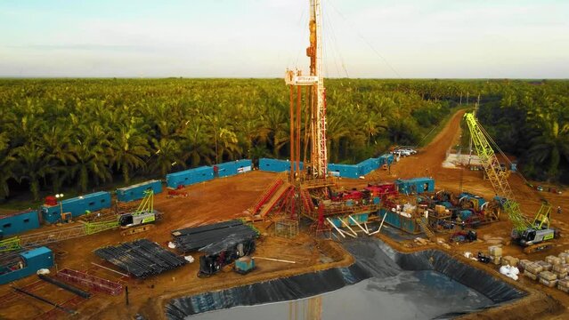 Cinematic 4K Drone Footage of Onshore Drilling Rig equipment structure for oil exploration and exploitation in the middle of jungle surrounded by palm oil trees during sunset and high oil price.