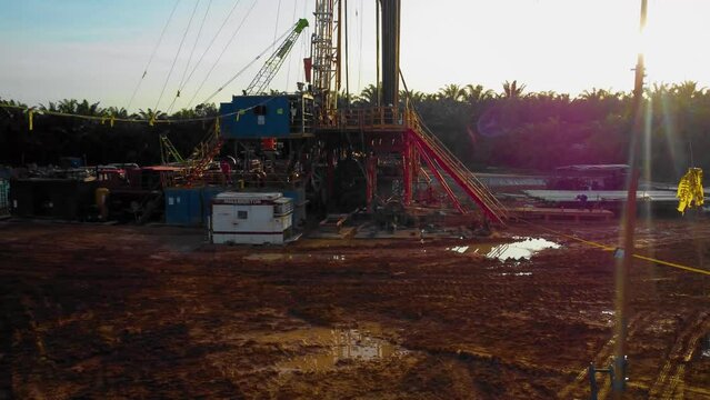 Cinematic 4K Drone Footage of Onshore Drilling Rig equipment structure for oil exploration and exploitation in the middle of jungle surrounded by palm oil trees during sunset and high oil price.