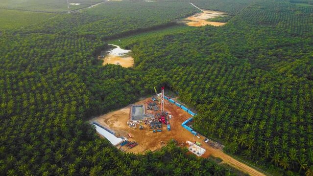 Cinematic 4K Drone Footage of Onshore Drilling Rig equipment structure for oil exploration and exploitation in the middle of jungle surrounded by palm oil trees during sunset and high oil price.