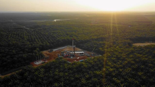 Cinematic 4K Drone Footage of Onshore Drilling Rig equipment structure for oil exploration and exploitation in the middle of jungle surrounded by palm oil trees during sunset and high oil price.
