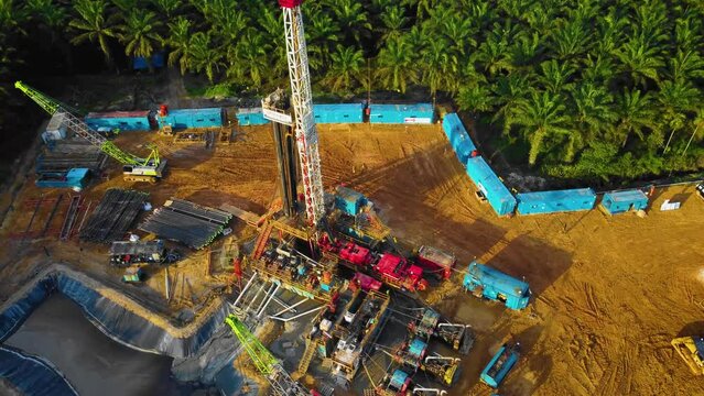Cinematic 4K Drone Footage of Onshore Drilling Rig equipment structure for oil exploration and exploitation in the middle of jungle surrounded by palm oil trees during sunset and high oil price.