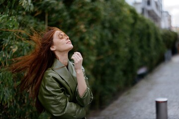 Fashion woman in the city on the street posing in front of buildings and trees with red long flyaway hair in the windy weather