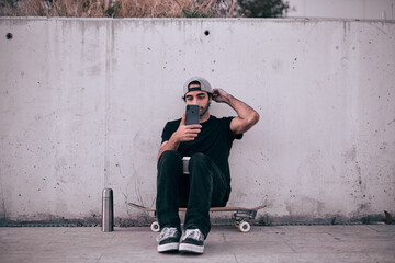 caucasian guy in gray cap and black t-shirt sitting on his skateboard manipulating his smartphone near the concrete wall