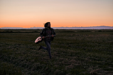 Caucasian young man with long hair in fur jacket running across the plain away from the mountains holding a guitar at sunset
