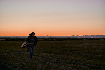 caucasian young man in fur jacket running across the big lonely green field next to his guitar at sunset