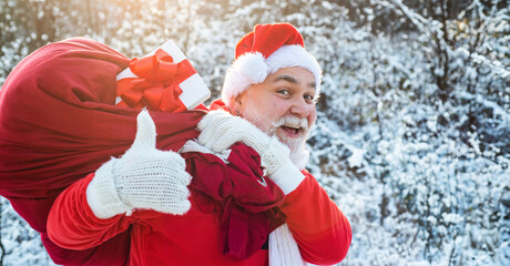 Obraz premium Santa claus with bag full of christmas presents outdoor. Santa portrait close up with thumbs up on winter snowy background. Santa Claus pulling huge bag of gifts on outside.