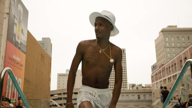 Attractive man wearing fedora hat and gold jewelry exits rooftop swimming pool dripping water as the camera tilts up at him in slow motion. Shot in 4k slow motion with skyline behind him.