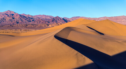Fototapeta na wymiar Sunset at Sand Dunes - Spring sunset view of smooth and curvy sand dunes towering at front of rugged purple mountains of Amargosa Range. Mesquite Flat Sand Dunes of Death Valley National Park, CA, USA