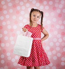 Sassy confident little girl in red polka dot dress holding gift bag on pink background - 548143958