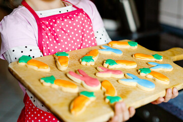 Closeup of cute little toddler girl holding fresh baked homemade Easter or spring cookies at home indoors. Adorable blond child with apron with bunny and carrot cookie in domestic kitchen.