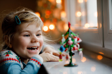 Litte toddler girl sitting by window and decorating small glass Christmas tree with tiny xmas toys. Happy healthy child celebrate family traditional holiday. Adorable baby.