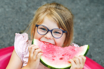 Cute little girl with glasses eating watermelon on inflatable ring in summertime. Happy smiling...