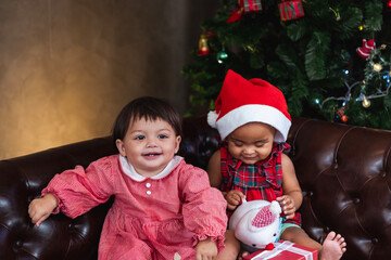 Two diverse baby friends are happily smiling while playing with present on couch dressing in christmas costume and santa hat with christmas tree on the back for season celebration concept