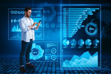 Attractive young european man using cellphone with abstract glowing blue business charts and graphs hologram on dark background. Innovation, future and fintech concept.