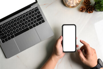 Top view of man hand using smartphone and laptop computer, lightbulb, pine cone on marble table