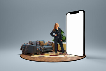 Young girl in homewear standing near to 3d model of cellphone with blank white screen and dreaming isolated on grey background. Online shopping, payment, new app or website - obrazy, fototapety, plakaty