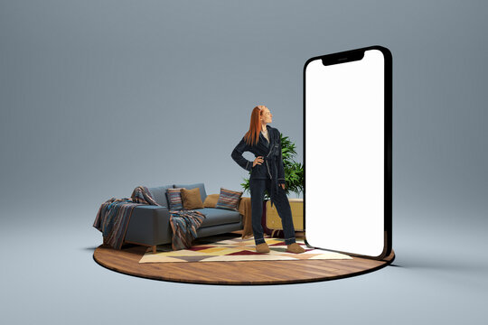 Young girl in homewear standing near to 3d model of cellphone with blank white screen and dreaming isolated on grey background. Online shopping, payment, new app or website