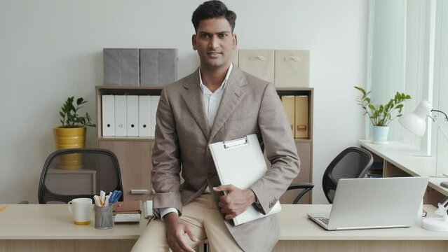 Tilt up shot of Indian businessman in formal suit sitting on desk in office, holding clipboard and posing for camera with smile