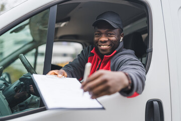 Positive postal package delivery. Friendly hard-working Black man in his 30s handling over a pen and delivery documents to the camera via driver's window. White delivery van. High quality photo