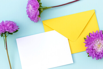 White blank card, yellow envelope and purple flower on blue background. Minimal style. Top view Flat lay Mockup