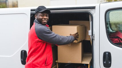 Upcoming holiday season sale via online shopping. Outdoor medium closeup shot of smiling surprised Black man in red-and-gray working clothes taking out one of many cardboard boxes out of his white van