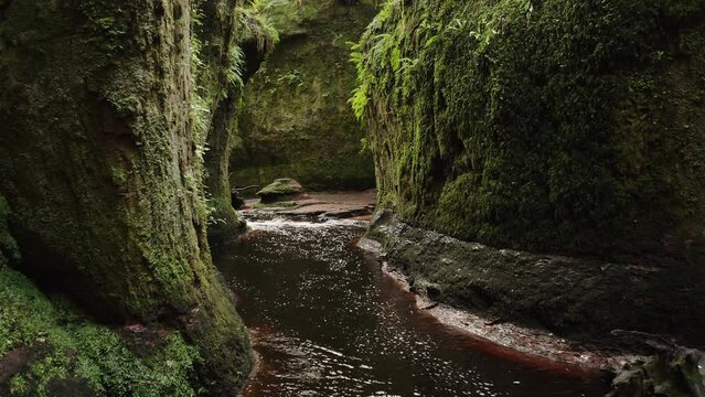 Drone shot creeping through the Devils Pulpit gorge in Scotland revealing mossy walls and a flowing red river.