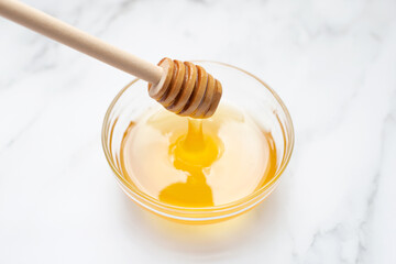 Honey flows from a wooden spoon into a glass bowl. Marble background.