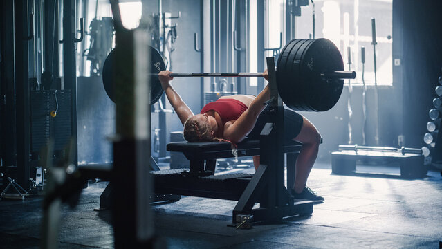 A Professional Bodybuilder Training by Lifting a Heavy Barbell In a Dark Gym. Female Energetic Athlete Doing Core Exercise with Bench Press Workout in an Empty Gym