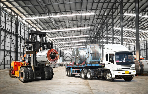 Forklift lifts heavy steel coil inside a warehouse at a seaport-bounded area to load onto truck. Raw material handling and transportation.
