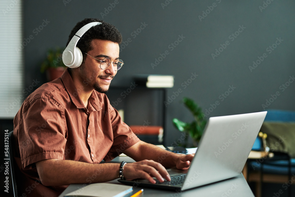 Poster young smiling man in headphones typing on laptop keyboard while sitting by workplace and taking part